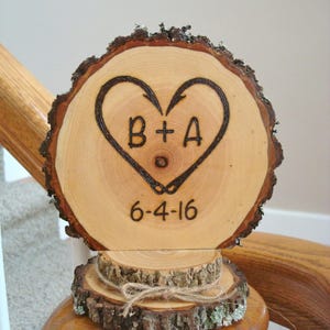 May include: A wooden heart-shaped wedding cake topper with the initials "B+A" and the date "6-4-16" burned into the wood. The topper is made of two slices of wood, one on top of the other.