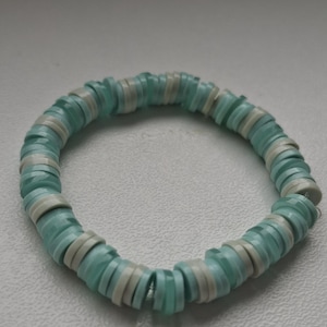 May include: A circular bracelet composed of alternating flat, disc-shaped beads in translucent light blue and opaque white. The beads are strung together in a repeating pattern, creating a continuous, flexible band with a smooth, uniform texture.