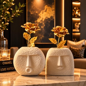 May include: Two off-white ceramic vases with textured surfaces, each shaped like a stylized human head with distinct facial expressions. Each vase holds a single metallic gold rose with detailed petals and leaves, creating a decorative display.