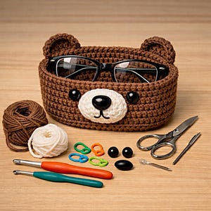May include: A brown crocheted bear-shaped basket holding black eyeglasses. The basket has a white muzzle and black button eyes. Knitting supplies, including yarn, hooks, and scissors, are arranged around the basket on a wooden surface.
