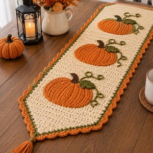 May include: A rectangular cream-colored crochet table runner featuring three orange pumpkin appliqués with green stems and vines. The runner has a scalloped orange border and a tassel at each end, showcasing a textured, handmade knit design.