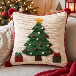 May include: A square crochet throw pillow with a cream-colored base, featuring a green Christmas tree with a yellow star, red and white ornaments, and two gift boxes at the base. The pillow has a red border and red pom-poms at each corner.