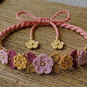 May include: A bracelet featuring a braided pink cord with an adjustable sliding knot closure. The band is decorated with small crocheted flowers in shades of pink, yellow, and dark red, accented with tiny gold-toned beads and two dangling flower charms.