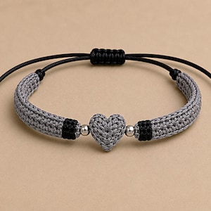 May include: A bracelet featuring a central gray crocheted heart, flanked by two silver-toned metallic beads and two black crocheted segments. The band is made of gray crocheted cord with black adjustable sliding knots at the ends for sizing.