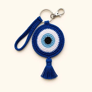 May include: A circular crocheted keychain featuring a concentric evil eye pattern in black, light blue, white, and dark blue yarn. The design includes a matching dark blue crocheted wrist strap, a silver metal keyring with a lobster clasp, and a tassel.