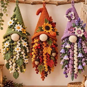Peut inclure: Trois décorations murales en crochet représentant des gnomes. Le gnome de gauche est vert avec des marguerites blanches. Celui du centre est orange avec des tournesols, des citrouilles et des feuilles d'automne. Celui de droite est violet.