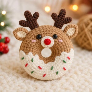 May include: A crocheted reindeer donut ornament featuring a brown body, white icing, and red, green, and pink sprinkles. The reindeer has dark brown antlers, two small ears, black bead eyes, and a bright red nose centered on the donut hole.