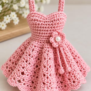 May include: A pink crochet dress featuring a sweetheart neckline, thin shoulder straps, and a flared skirt with a textured pattern. A small crocheted flower with a central white pearl bead is attached to the waist, with two dangling cords.