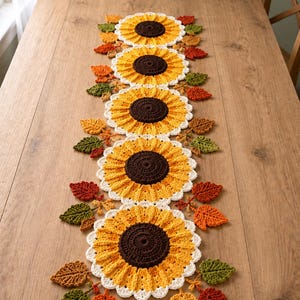 May include: A long, rectangular crochet table runner featuring five large sunflower motifs with dark brown centers, bright yellow petals, and white scalloped edges, accented by small, textured autumn leaves in shades of orange, red, and green.