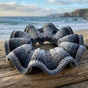 May include: A handmade, gray and white crochet scrunchie with a wavy design. The scrunchie is resting on a piece of driftwood on a beach, with the ocean and coastline in the background. The scrunchie is approximately 6 inches in diameter.