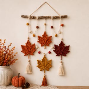 May include: A wall hanging featuring four crocheted maple leaves in shades of orange, rust, and yellow, suspended from a natural wooden branch by cream-colored macrame cord. The design includes small wooden beads, pom-poms, and cream-colored tassels.