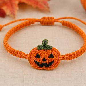 May include: An orange macrame bracelet featuring a central crocheted pumpkin charm. The pumpkin has a dark green stem and a black stitched jack-o'-lantern face. The bracelet band is made of orange cord with an adjustable sliding knot closure.