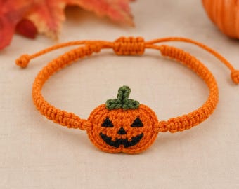 May include: An orange macrame bracelet featuring a central crocheted pumpkin charm. The pumpkin has a dark green stem and a black stitched jack-o'-lantern face. The bracelet band is made of orange cord with an adjustable sliding knot closure.