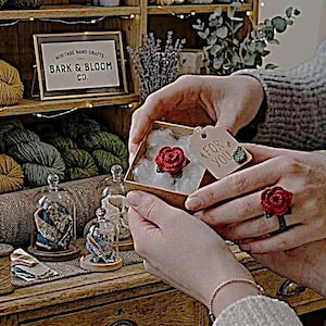 May include: A person holding a small box with a red rose inside and a matching rose ring. The box has a "FOR YOU" tag. The background includes shelves of yarn, decorative glass domes, and a sign that reads "BARK & BLOOM CO."