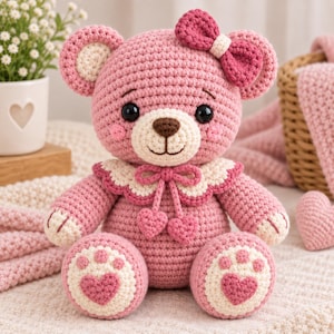 May include: A pink crocheted teddy bear featuring a cream-colored muzzle, inner ears, and paw pads. The bear wears a cream-colored scalloped collar with two small heart-shaped charms and a matching pink bow on its head. It has black eyes.