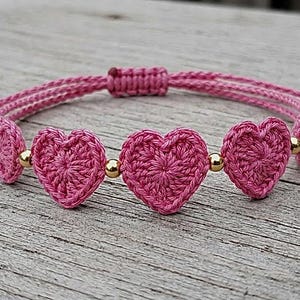 May include: A pink crochet heart bracelet featuring five small, textured heart-shaped motifs linked by polished gold-tone metal beads. The bracelet has a braided cord band with an adjustable sliding knot closure and gold-tone beads at the ends.