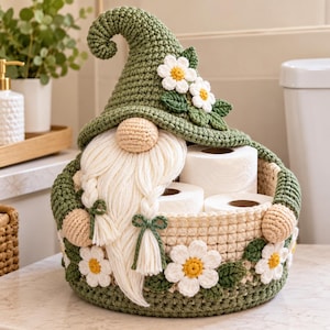 May include: A crochet gnome-shaped toilet paper basket in olive green and cream. The gnome features a tall pointed hat with white daisy appliqués, a round beige nose, and a long cream-colored yarn beard with two braids tied with green bows.