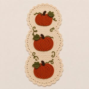Puede incluir: Un camino de mesa de ganchillo color crema con tres secciones circulares, cada una decorada con una calabaza naranja texturizada, un tallo marrón oscuro, una hoja verde y pequeñas enredaderas verdes, con un borde festoneado.