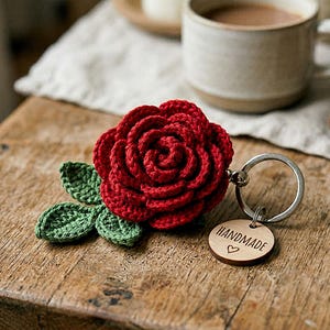 May include: A handmade red crochet rose keyring with green leaves and a wooden tag that says "HANDMADE" with a heart. The keyring is on a wooden surface.
