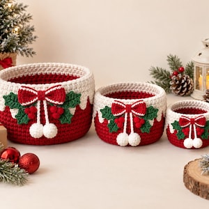 Puede incluir: Tres cestas de almacenamiento redondas de ganchillo en tamaños descendentes, con una base de color rojo intenso y un borde superior crema. Cada cesta está decorada con un lazo rojo, hojas de acebo verdes, bayas rojas y dos pompones blancos.