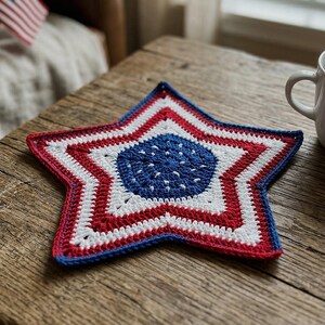 May include: A handcrafted, star-shaped crochet coaster in red, white, and blue, reminiscent of the American flag. The coaster is placed on a rustic wooden table, next to a white mug. The coaster's design features a blue center with white stars.