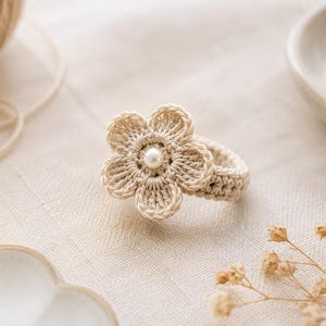 Puede incluir: Un anillo de ganchillo en color beige con un diseño de flor de seis pétalos y una pequeña perla blanca en el centro. La banda del anillo está hecha de hilo beige a juego, mostrando una apariencia texturizada y cosida a mano.