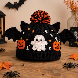 Op de afbeelding: Een zwarte gehaakte muts met een witte spookapplicatie, twee oranje pompoenapplicaties en zwarte vleermuisvleugels aan de zijkanten. De muts is afgewerkt met een oranje en zwarte pompon en versierd met kleine witte stersteekjes.