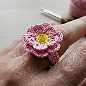 Peut inclure: Une bague faite à la main en crochet en forme de fleur rose avec un centre jaune. La bague est portée au doigt, mettant en valeur les détails complexes du motif floral. Une pelote de fil rose et un crochet sont en arrière-plan.