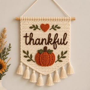 May include: A cream-colored crocheted wall hanging featuring the word thankful in dark brown yarn, a burnt orange pumpkin, and a small heart above the text. The piece is suspended from a wooden dowel with a white cord and has eight tassels.