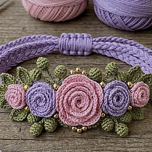 May include: A handcrafted crochet bracelet featuring a central arrangement of pink and lavender roses, accented with green crocheted leaves and small gold-toned beads. The band is made of braided lavender thread with an adjustable sliding knot closure.