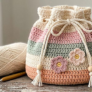 May include: A crochet drawstring bag featuring horizontal stripes in cream, dusty pink, sage green, and peach. The bag is adorned with two small crocheted flowers and includes a cream-colored drawstring cord with wooden beads and tassels at the ends.