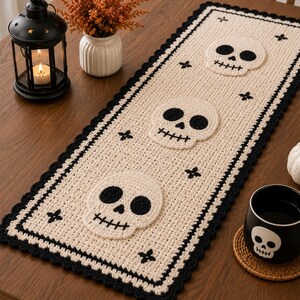 Peut inclure: Un chemin de table rectangulaire en crochet couleur crème avec une bordure festonnée noire. Le chemin présente trois motifs de crânes blancs centrés avec des orbites noires et des bouches cousues, agrémentés de petites croix noires.