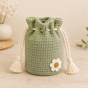 Può includere: Una pochette a sacchetto in crochet color verde salvia con motivo a maglia testurizzato e un'applicazione a forma di margherita bianca con centro giallo. La borsa presenta due cordini intrecciati color crema con nodi e nappe decorative.