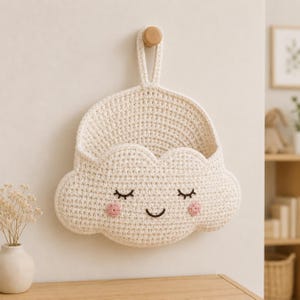 Puede incluir: Una cesta de pared de ganchillo color crema con forma de nube y cara sonriente. La cesta presenta ojos cerrados bordados, una pequeña boca curvada y dos mejillas circulares rosas. Incluye un asa única en la parte superior para colgarla.