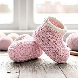 以下が含まれることがあります： 白のトリムが付いた、手作りのピンク色のベビー用かぎ針編みブーツ2足。ブーツは、背景に毛糸玉がある、風化した木製の表面に展示されています。かぎ針編みのステッチが見え、アイテムの職人技を示しています。