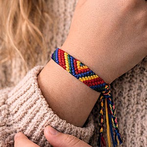 May include: A woven friendship bracelet featuring a chevron pattern in vibrant red, yellow, green, and blue threads. The bracelet is tied with braided cord ends that hang down, showcasing a multi-colored design crafted from thin, durable textile strands.