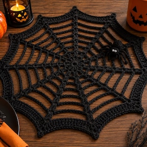 May include: A black crocheted spider web doily featuring a small, attached black spider with white eyes. The web has a circular center with radiating spokes and concentric rings, creating a detailed, open-work geometric pattern in black yarn.