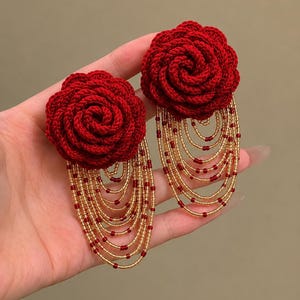 May include: A pair of earrings featuring deep red crocheted rose flowers. Attached to the base of each flower are multiple cascading strands of gold-toned seed beads interspersed with small red beads, creating a layered fringe effect.