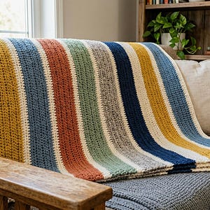 Puede incluir: Una manta rectangular de ganchillo con rayas verticales en color crema, amarillo mostaza, azul marino, naranja quemado, verde salvia y gris jaspeado. La manta presenta un patrón de punto texturizado y está rematada con flecos color crema.