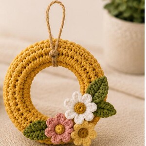 Könnte beinhalten: Ein kreisförmiger, gelber Häkelkranz mit einer Aufhängeschlaufe aus Jute. Der Kranz ist mit drei kleinen gehäkelten Blumen in Weiß, Rosa und Gelb sowie zwei grünen gehäkelten Blättern an der unteren rechten Seite verziert.