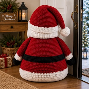 May include: A large, round, crocheted Santa Claus figure featuring a red hat with a white pom-pom, a white brim, a red body, a black belt, and white trim at the base. The figure is crafted with a textured yarn stitch and has a rounded, plush shape.