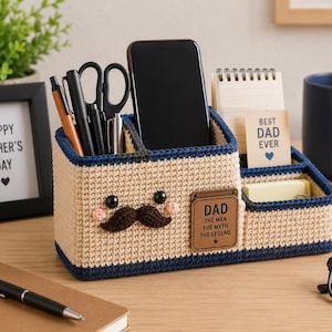 May include: A beige and navy blue crochet desk organizer featuring a mustache, two black eyes, and pink cheeks. A brown leather-like tag is attached with the text DAD THE MAN THE MYTH THE LEGEND. The organizer holds a smartphone, pens, and a notepad.