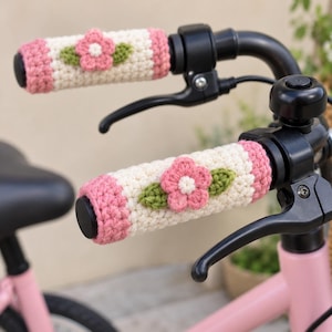 Könnte beinhalten: Ein Paar gehäkelte Fahrradlenkergriffe mit einer cremefarbenen Basis und rosafarbenen Enden. Jeder Griff ist mit einer dreidimensionalen rosa Blume mit weißer Mitte und zwei grünen Blättern in der Mitte des cremefarbenen Bereichs verziert.