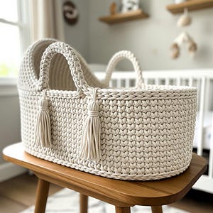 Puede incluir: Cuna de bebé de ganchillo color crema, con dos asas trenzadas y borlas decorativas. La cesta de forma ovalada se apoya sobre un soporte de madera. La decoración de la guardería es neutra y suave, con un enfoque en materiales naturales.