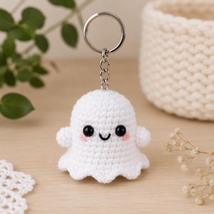 May include: A white crocheted ghost keyring featuring a rounded head, two small arms, and a scalloped bottom edge. The ghost has two black plastic safety eyes, a small embroidered black smile, and two light pink embroidered blush marks.