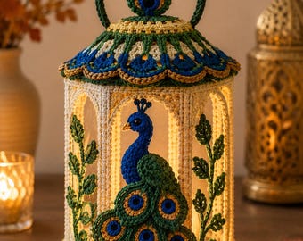 May include: A hexagonal lantern featuring intricate crochet work in cream, green, blue, and gold. The structure includes a peacock motif with a fanned tail, leafy vine accents on the side panels, and a decorative domed top with a circular handle.