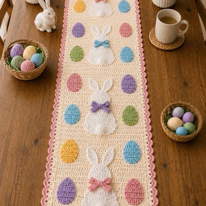 May include: A rectangular cream-colored crochet table runner featuring four white bunny motifs with colorful bows and alternating pastel-colored crocheted egg shapes. The runner has a scalloped pink border and is displayed on a wooden table.