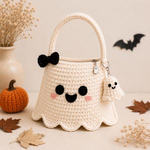 May include: A cream-colored crochet handbag shaped like a ghost with a scalloped bottom edge. It features a black crochet bow, two black circular eyes, a black smiling mouth, and two pink cheeks. A small ghost charm and a crochet ghost keychain are attached.