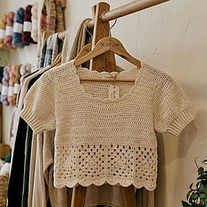 Puede incluir: Top de crochet color crema de manga corta con escote y dobladillo festoneados, colgado de una percha de madera. El top tiene un patrón calado decorativo en la cintura. The Cozy Loop está impreso en la percha. Se ve ropa y lana.