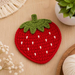 Puede incluir: Un posavasos con forma de fresa hecho de hilo rojo con un tallo y hojas verdes de ganchillo en la parte superior. El cuerpo rojo presenta pequeñas puntadas blancas bordadas dispersas por la superficie para representar las semillas.